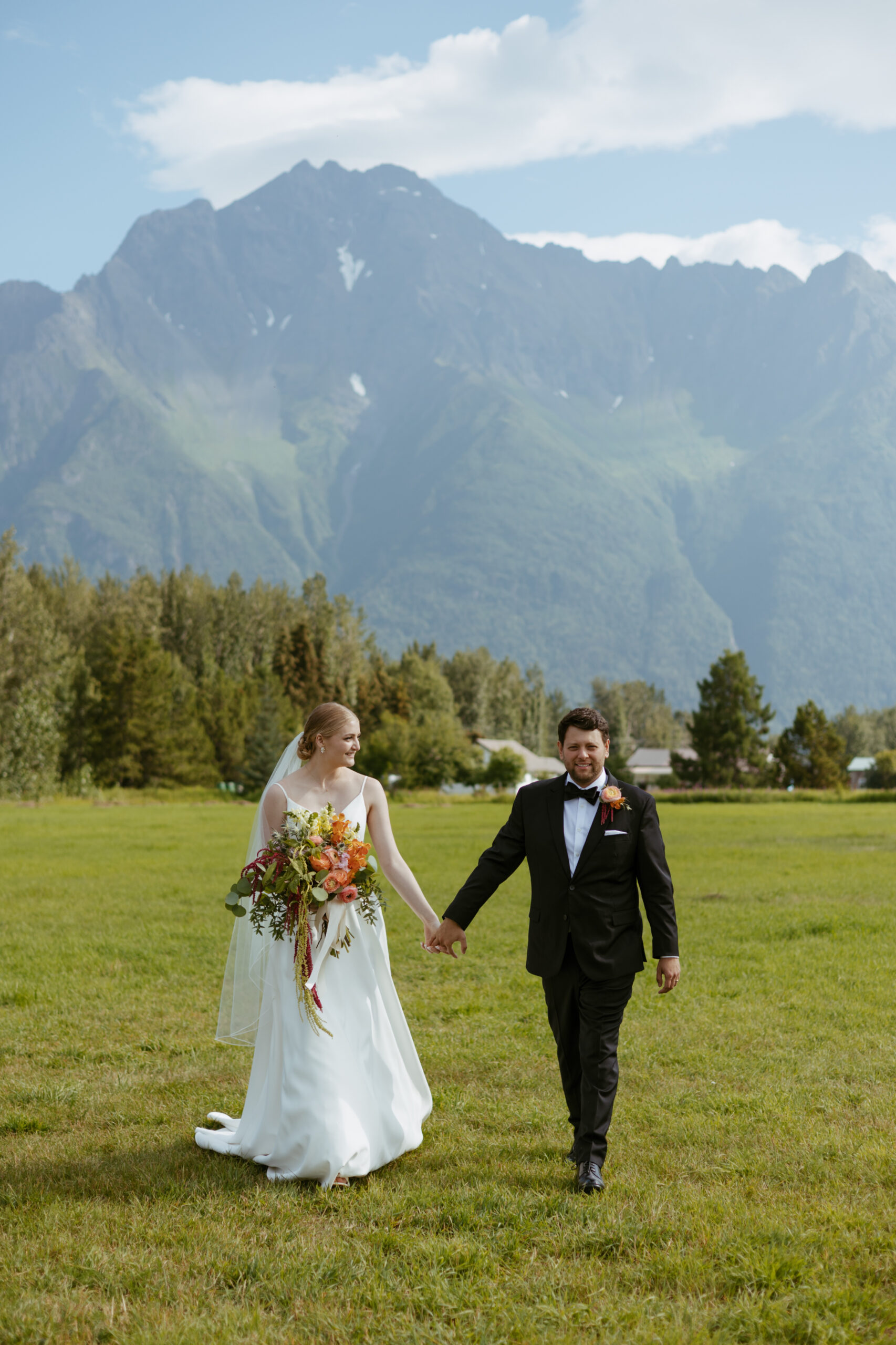 Wedding couple holding hands while walking together in a field with a mountain in the background.