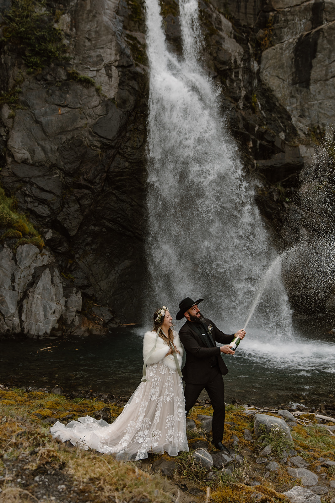 A couple who is in wedding attire sprays champagne together. They are standing in front of a waterfall