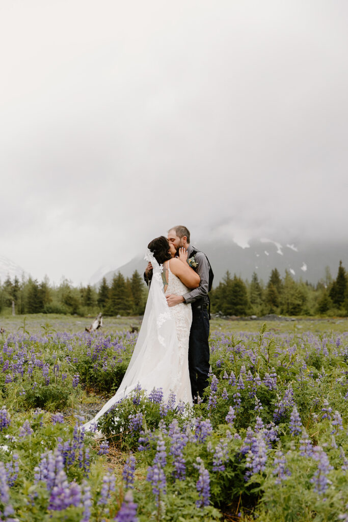 Bride and groom sharing their first kiss as newlyweds on a misty day in a field of purple lupine flowers, with mountains in the background.