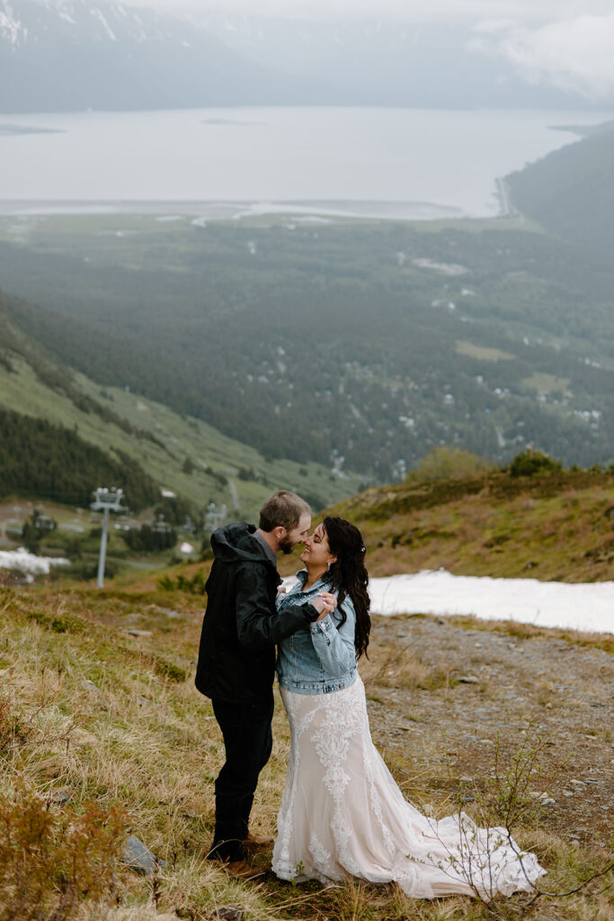Newlyweds smiling and embracing with interlocked fingers at the top of Alyeska Mountain in Girdwood, Alaska, with Turnagain Arm visible in the distance.