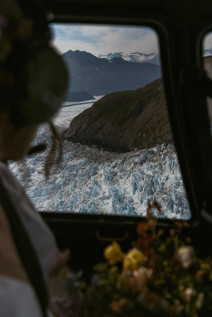 Bride wearing a headset looking out the helicopter window at an aerial view of Knik Glacier in Alaska, captured from inside the aircraft.