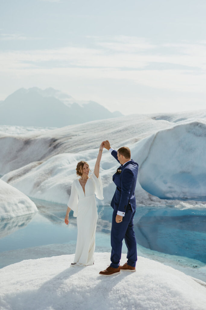 Bride and groom twirling joyfully on Knik Glacier in Alaska on a sunny day, with a turquoise glacier pool behind them.