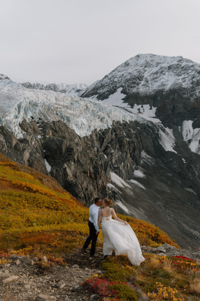 Bride and groom standing together on a mountain overlooking Knik Glacier in Alaska, surrounded by vibrant fall colors on the tundra.