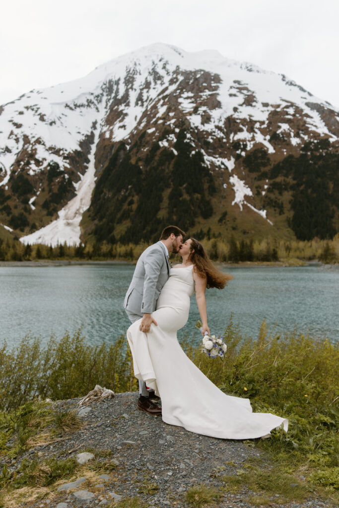 Groom dipping and kissing his bride at a scenic overlook with mountains and a lake in the background.