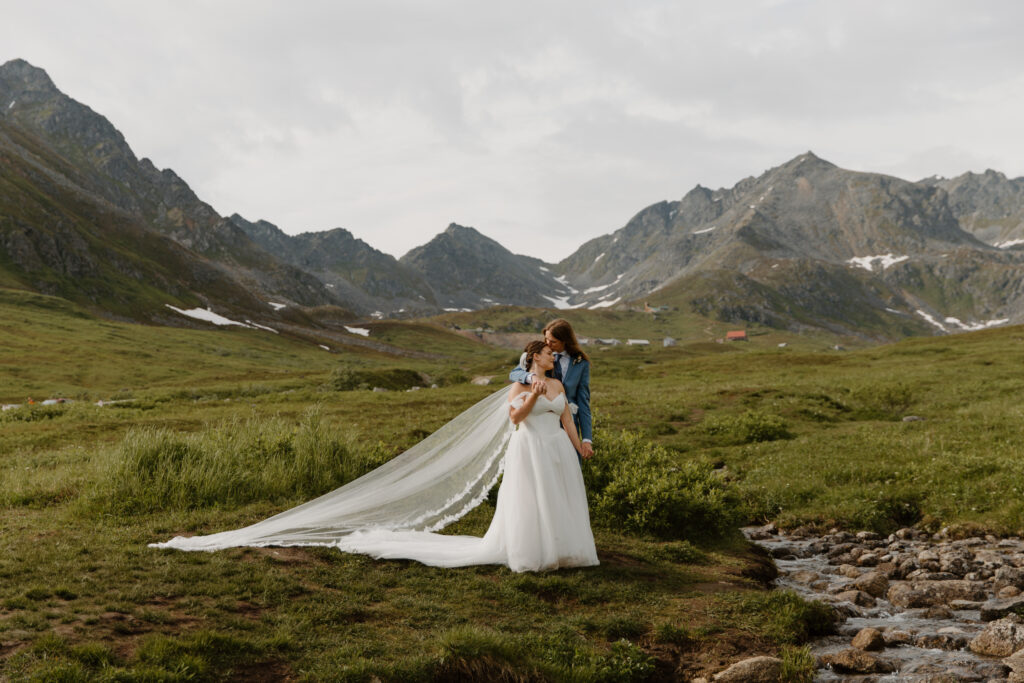 Bride and groom embracing in lush, green Hatcher Pass, Alaska, with a creek and majestic mountains in the background.
