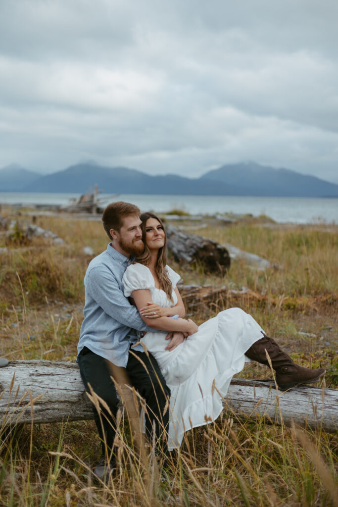 Engaged couple embracing while sitting on large driftwood on the beach in Homer, Alaska.