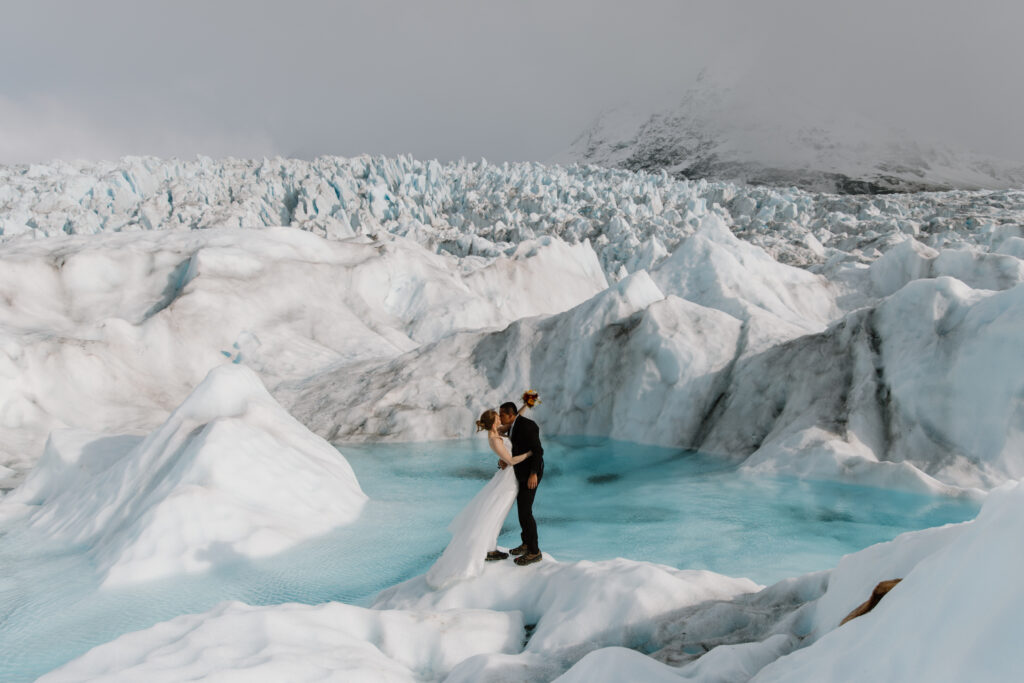 Newlywed couple embracing and kissing on Knik Glacier in Alaska, bride raising bouquet triumphantly, surrounded by jagged ice peaks.
