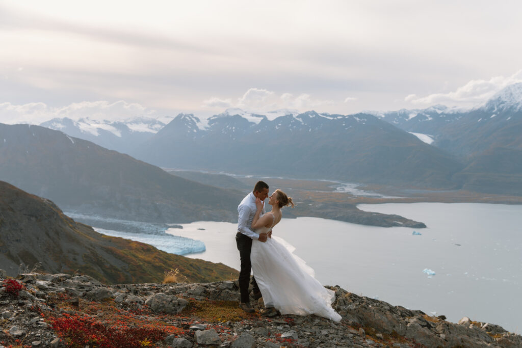 Bride and groom smiling forehead-to-forehead on mountainside, embracing with distant mountains and lake in the valley below.