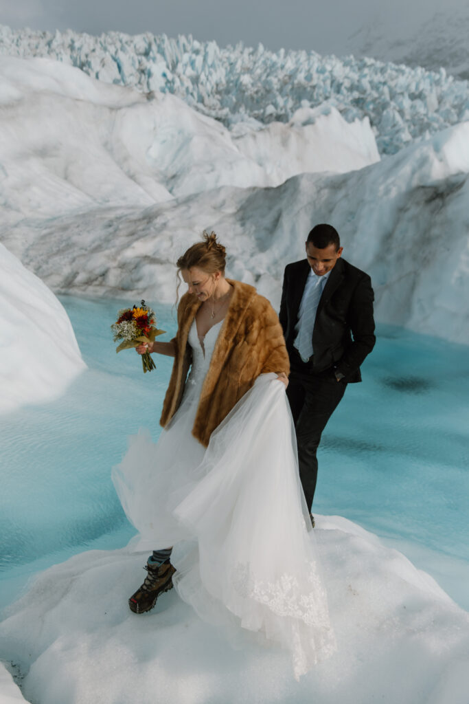 Newlywed couple smiling and walking hand in hand on Knik Glacier ice after ceremony, bride wearing fur shawl for warmth.