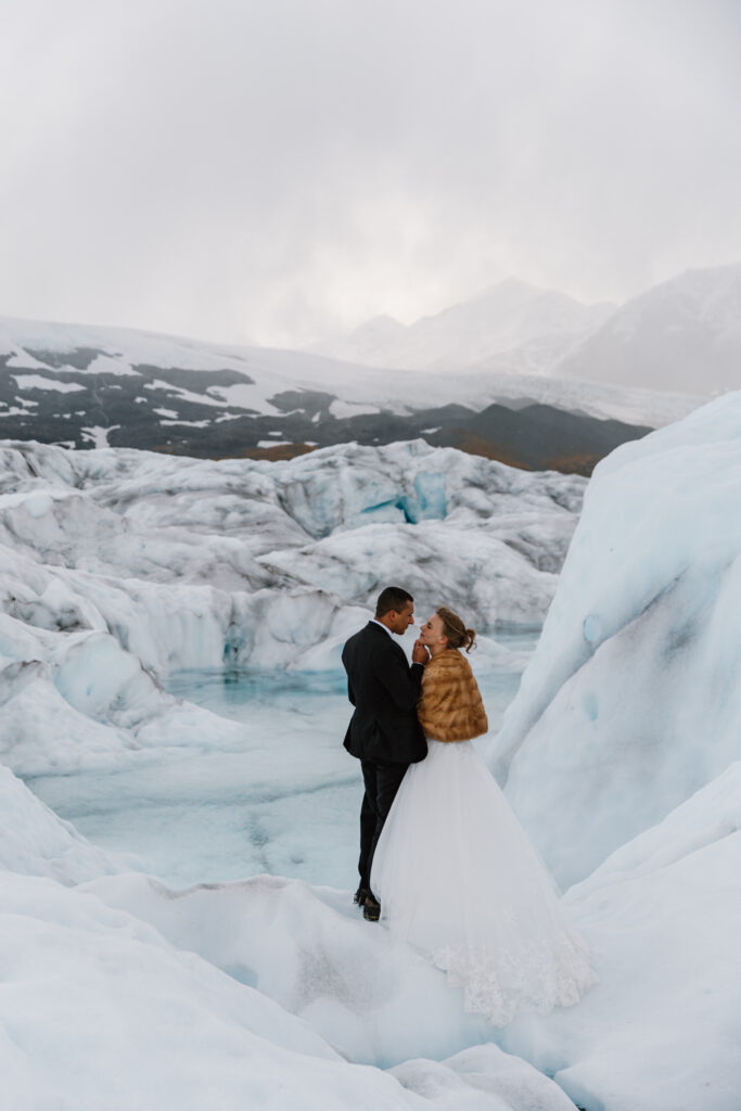 Bride and groom standing on Knik Glacier, groom gently lifting bride's chin as they gaze at each other during portraits.