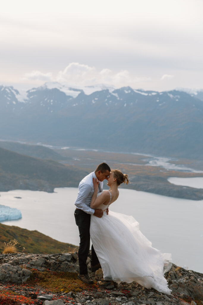 Bride and groom smiling forehead-to-forehead on mountainside, embracing with distant mountains and lake in valley below.
