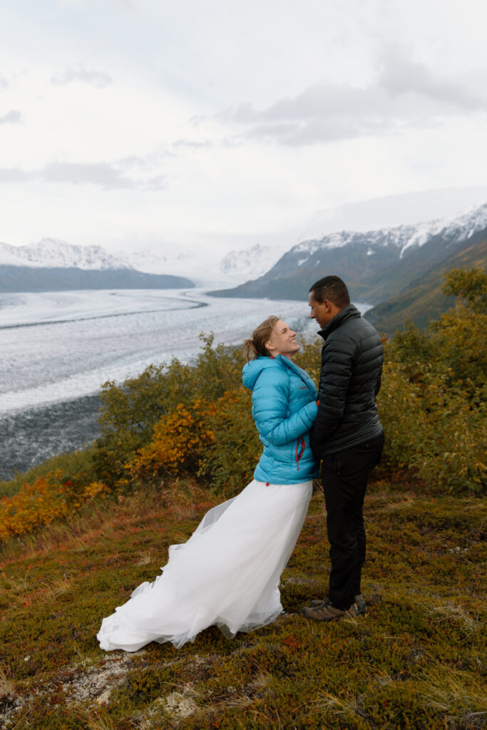 Bride and groom in puffy jackets (bride in blue) smiling at each other, with Knik Glacier and mountains visible in the distance.