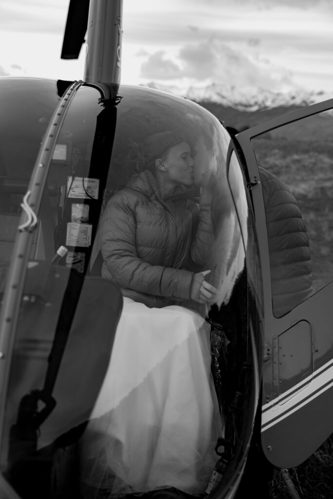 Black and white photo of bride sitting in helicopter while groom stands outside door kissing her before glacier elopement takeoff.