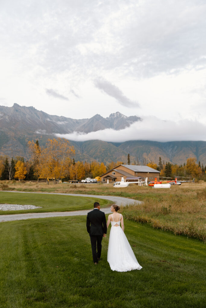 Bride and groom walking hand in hand on green lawn toward helicopters, smiling at each other with Palmer mountains in the distance.