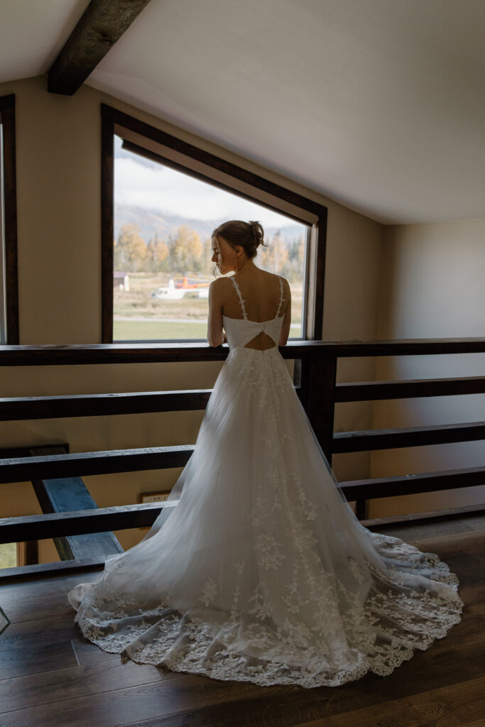 Bride standing on second-floor balcony of cabin, back to camera, gazing to the side with Palmer Valley mountain views in Alaska.