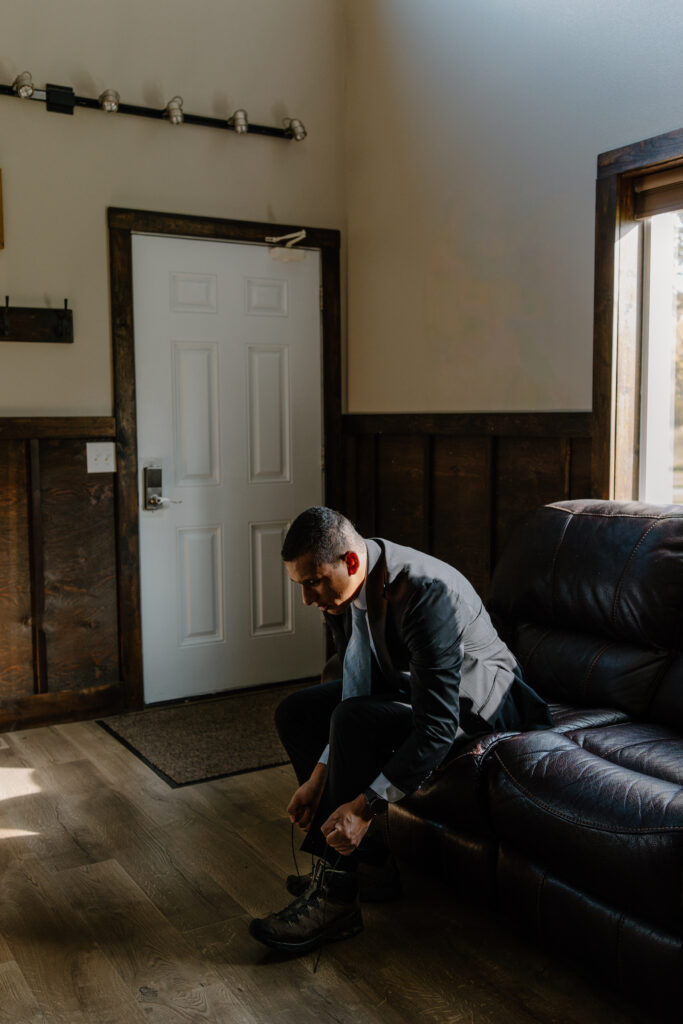Groom sitting on couch in cabin, lacing up his shoes during getting-ready moments before glacier elopement.
