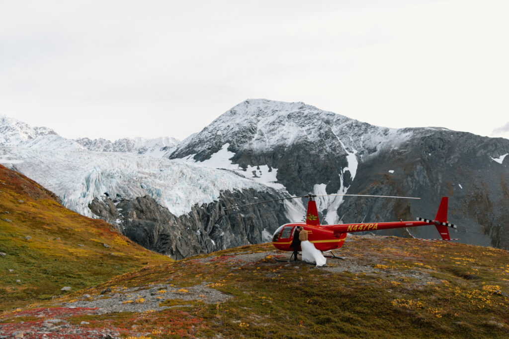 Bride and groom loading into a red helicopter on a mountainside near Knik Glacier, same colorful fall tundra backdrop.