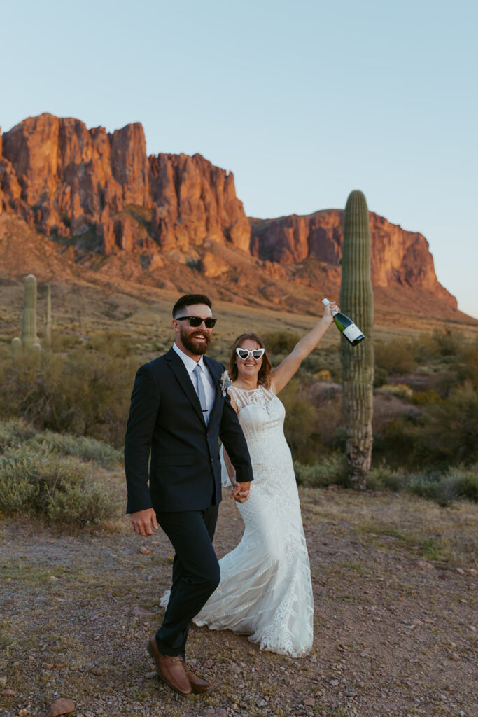 Newlywed couple walking hand in hand along a trail at Lost Dutchman State Park, both wearing sunglasses. The bride raises a champagne bottle triumphantly in celebration, with Superstition Mountains and green desert landscape in the background at sunset.