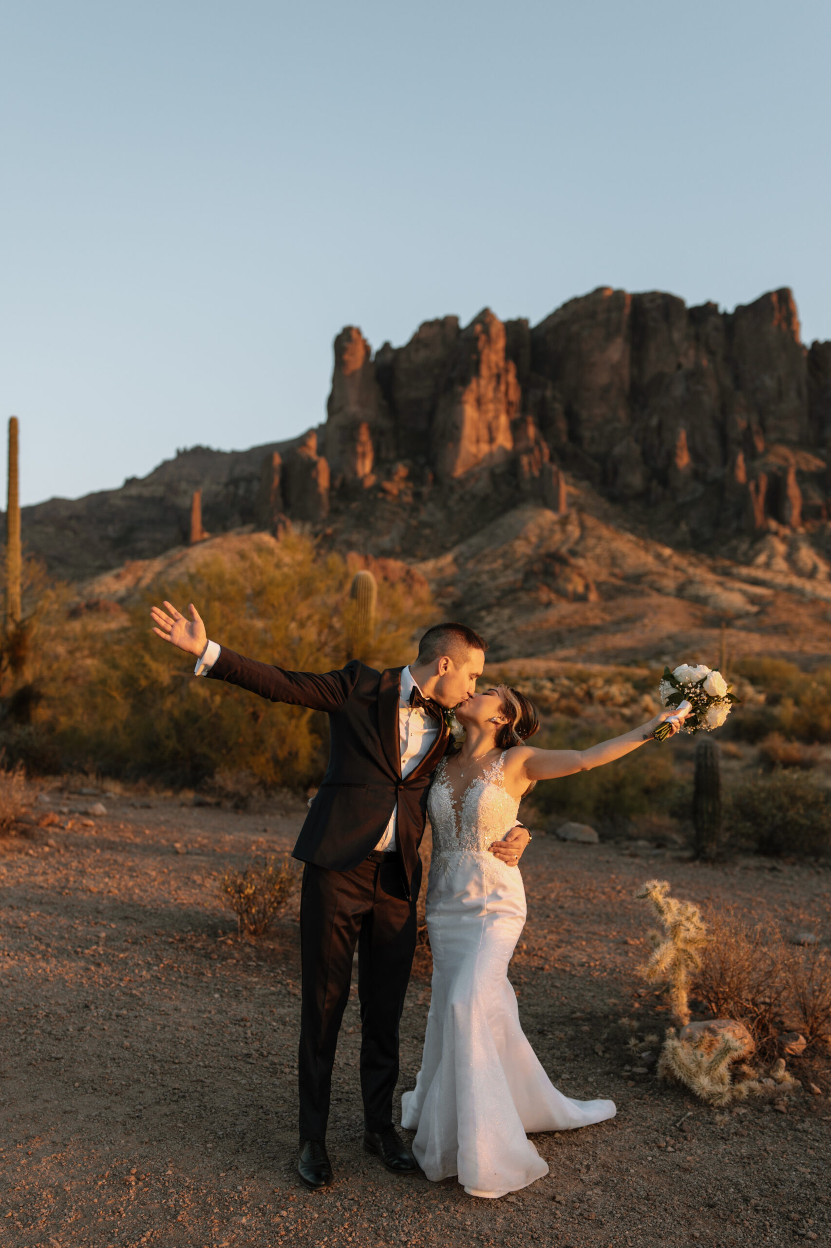 Bride and Groom raise their hands as the lean in to kiss each other while at sunset in Lost Dutchman State Park.