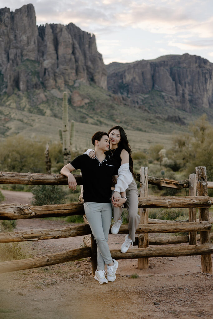 Asian couple leaning on each other in front of Superstition Mountains backdrop at Lost Dutchman State Park. The boyfriend leans against a rustic wooden fence while the girlfriend sits on it, both in casual clothes during golden hour sunset.