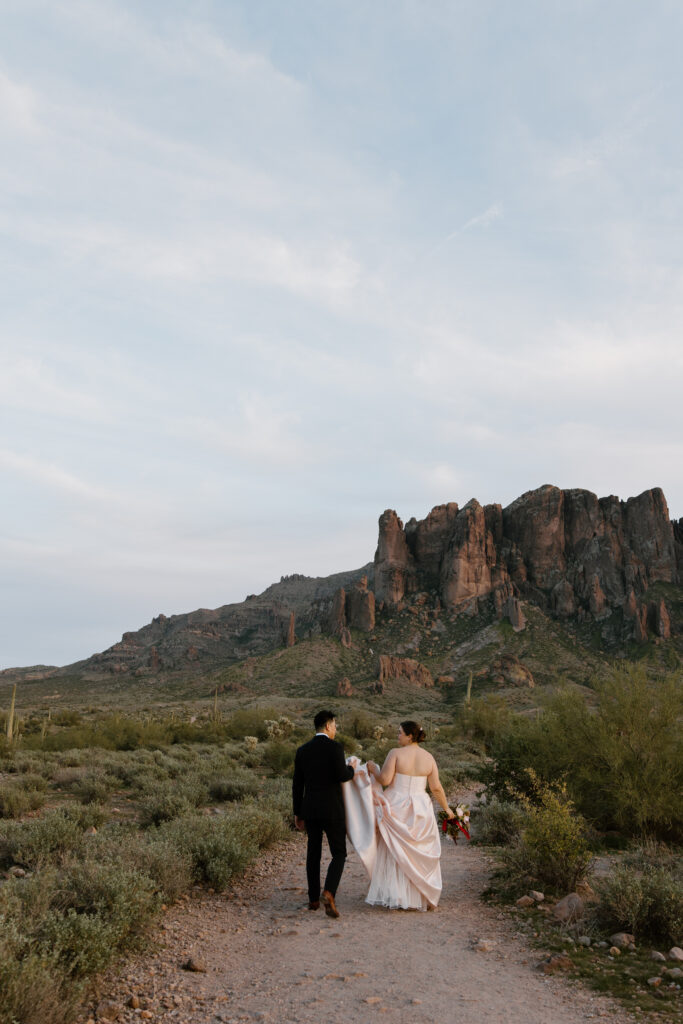 Newlywed couple walking hand in hand on a trail at Lost Dutchman State Park. Groom gently holds the bottom of the bride's dress as they talk and smile, with Superstition Mountains and desert landscape in the background.