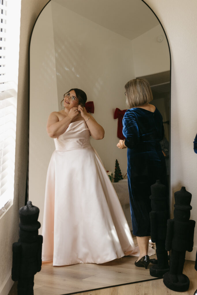 Bride putting on earrings in front of a full-length mirror in a home, with her mom standing beside her offering support during getting-ready.
