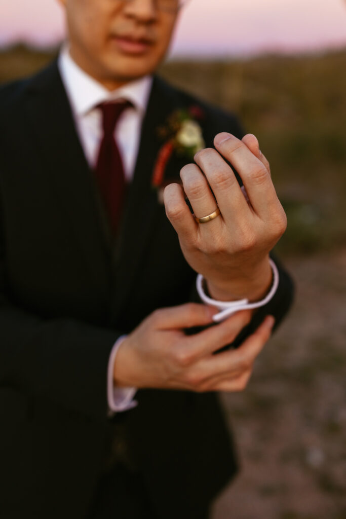 Close-up of groom buttoning his shirt cuffs at sunset in Lost Dutchman State Park, with his wedding ring visible on his finger.