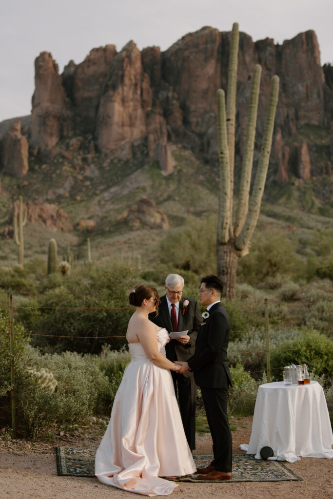 Wedding couple holding hands at the altar on a rug, with the bride's father officiating the ceremony at Lost Dutchman State Park. Superstition Mountains rise dramatically in the background.