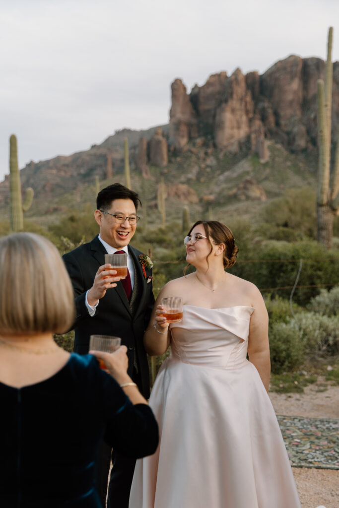 Newlywed couple cheering whiskey glasses with guests after toasts at Lost Dutchman State Park. The bride smiles warmly at her husband while raising her glass, with Superstition Mountains and desert landscape in the background.