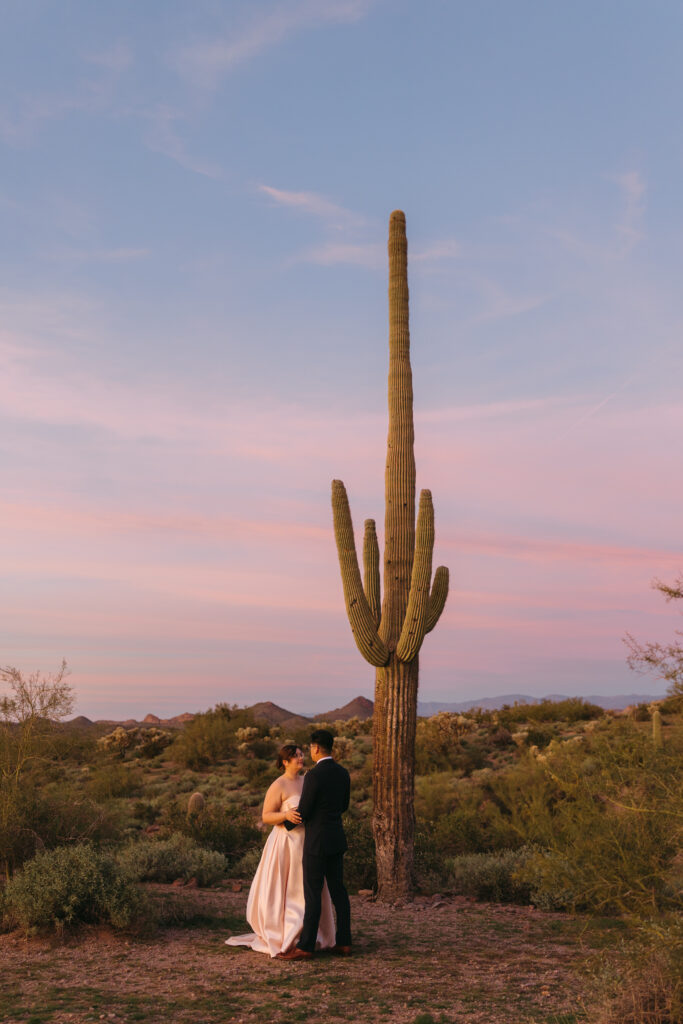 Newlywed couple embracing and gazing at each other beside a towering saguaro cactus at Lost Dutchman State Park during blue hour. The sky glows with vivid pink and purple colors as the Superstition Mountains fade into twilight.