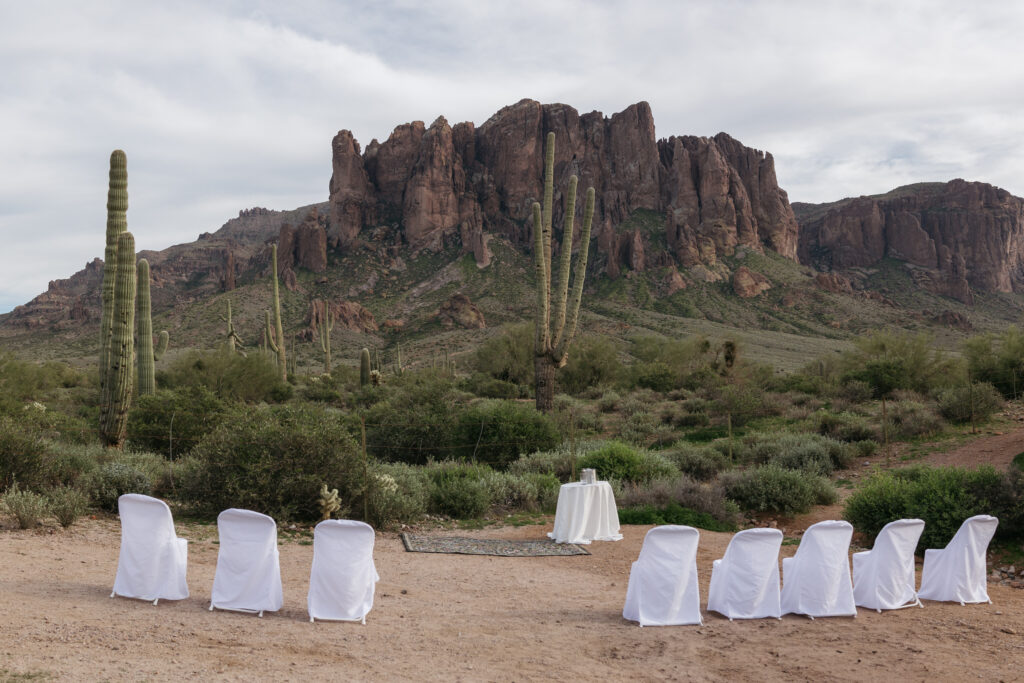 Empty ceremony setup at Lost Dutchman State Park: white chairs and rug arranged on a trail with Superstition Mountains backdrop at sunset, surrounded by green desert landscape. Photo by Brisa Breeze Photo.