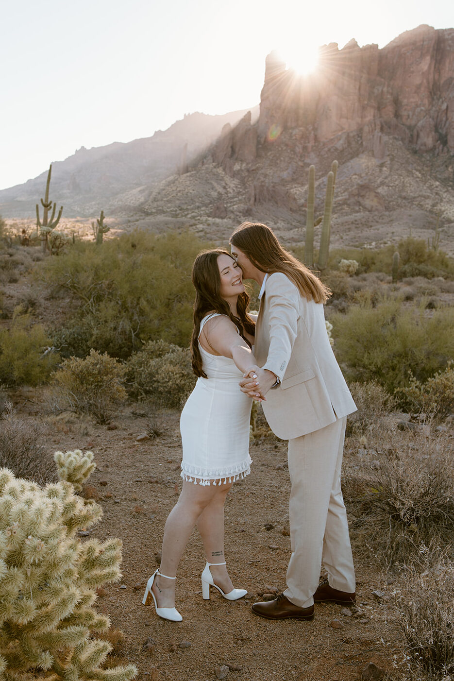 Engaged couple facing each other with hands clasped in front of Superstition Mountains backdrop at Lost Dutchman State Park. The guy kisses his fiance's cheek while she smiles with eyes closed, wearing a white short dress and he in a tan casual suit during sunrise.