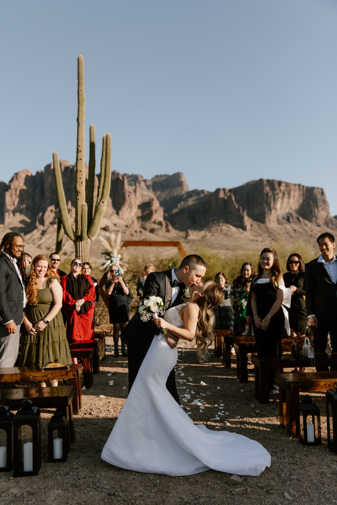 Newlywed couple sharing a dip kiss at the end of the altar after their ceremony at Lost Dutchman State Park. Guests cheer on both sides, with Superstition Mountains and green desert landscape in the background.