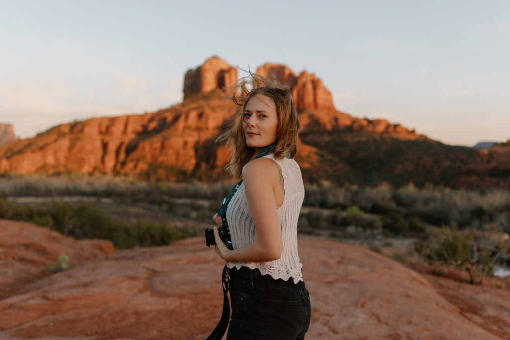 Photographer Brisa Breeze holding a film camera in front of Cathedral Rock at sunset in Sedona, Arizona. Wind blows through her hair as she turns back to look over her shoulder, with the iconic red rock formations glowing in golden-hour light.
