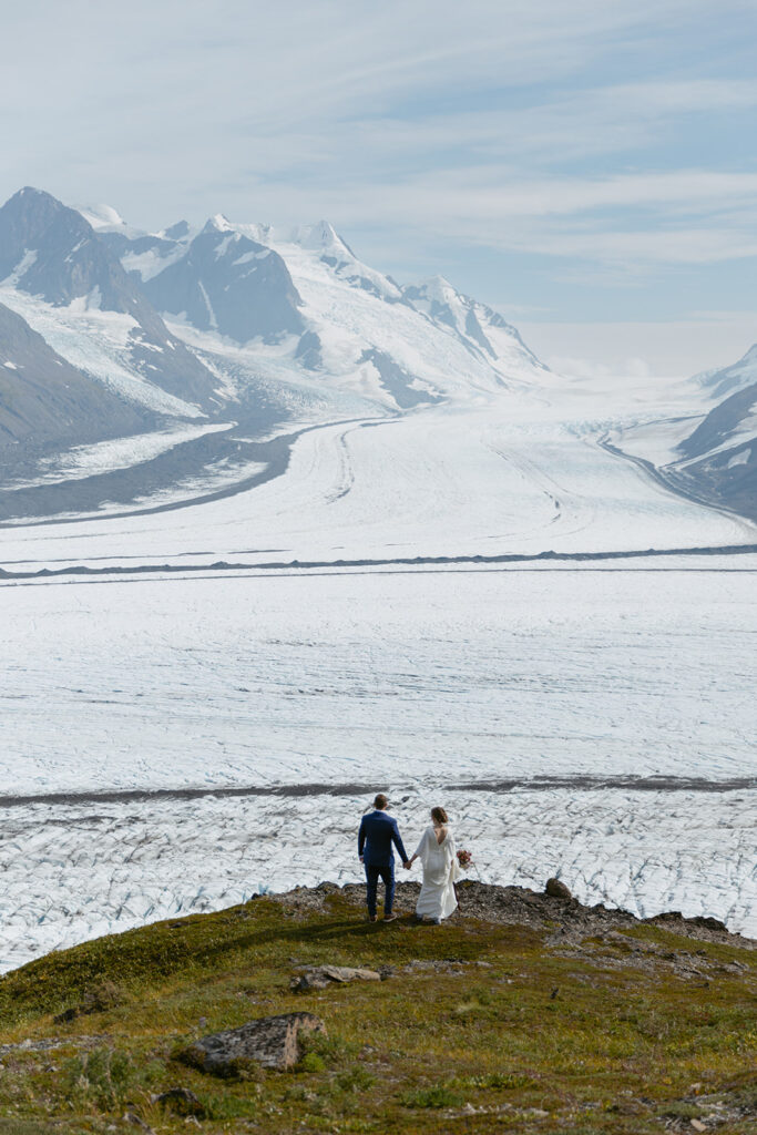 Bride and groom walking to the edge of an overlook with a view of Knik Glacier on a sunny day in Alaska