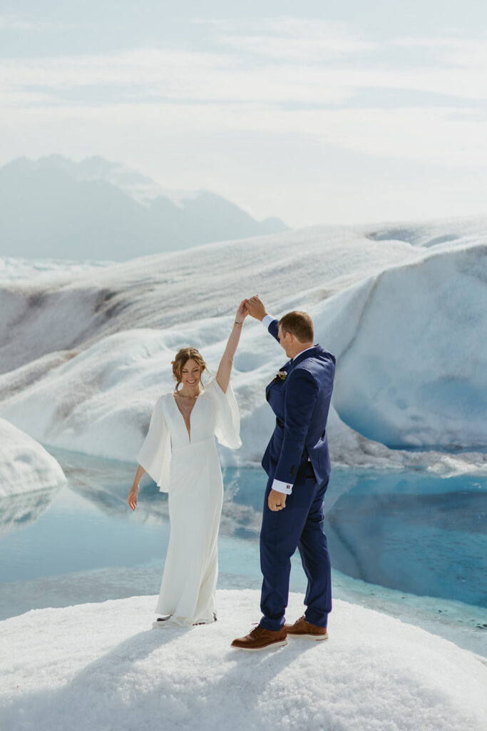 Groom twirling bride on Knik Glacier with a glacial pool visible behind them in Alaska