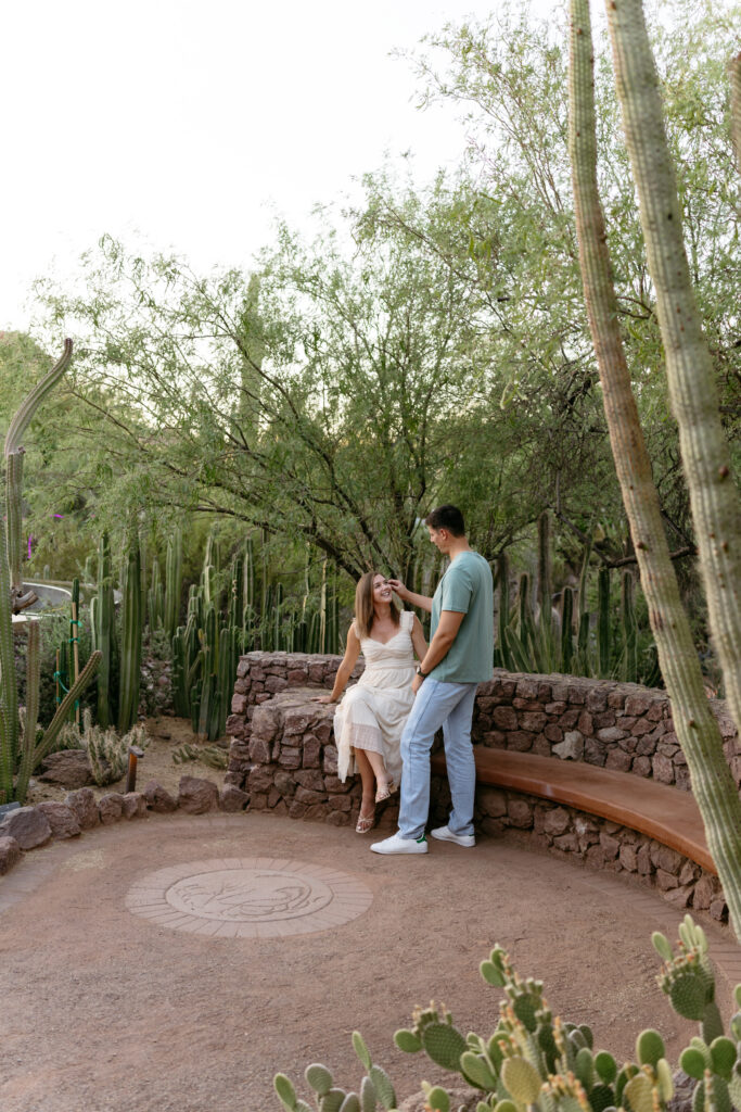 Engaged couple sit on a bench with cacti behind them at the Phoenix Botanical Garden