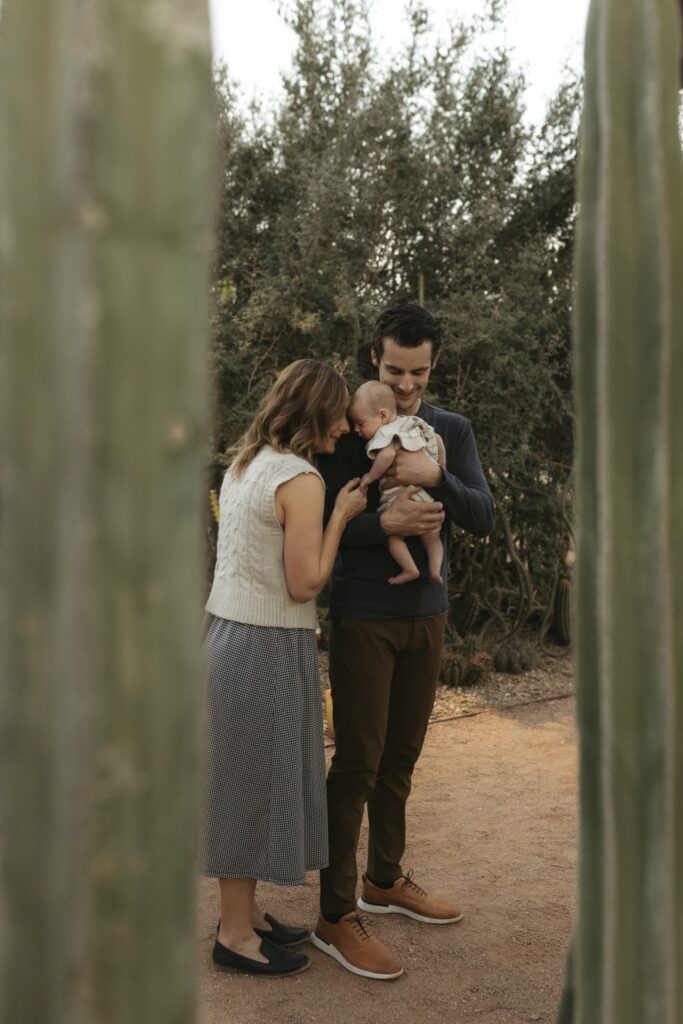 Mom, dad, and baby are standing and cradling their newborn. They are framed back cactus on either side,
