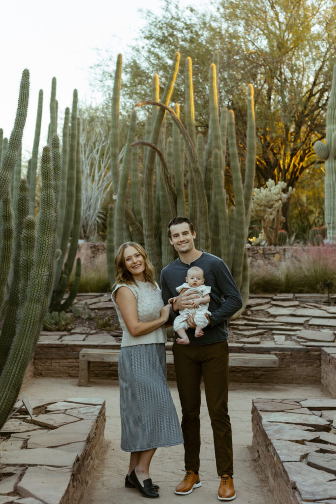 Mom, dad, and baby are smiling at camera. There are tall cactus behind them.
