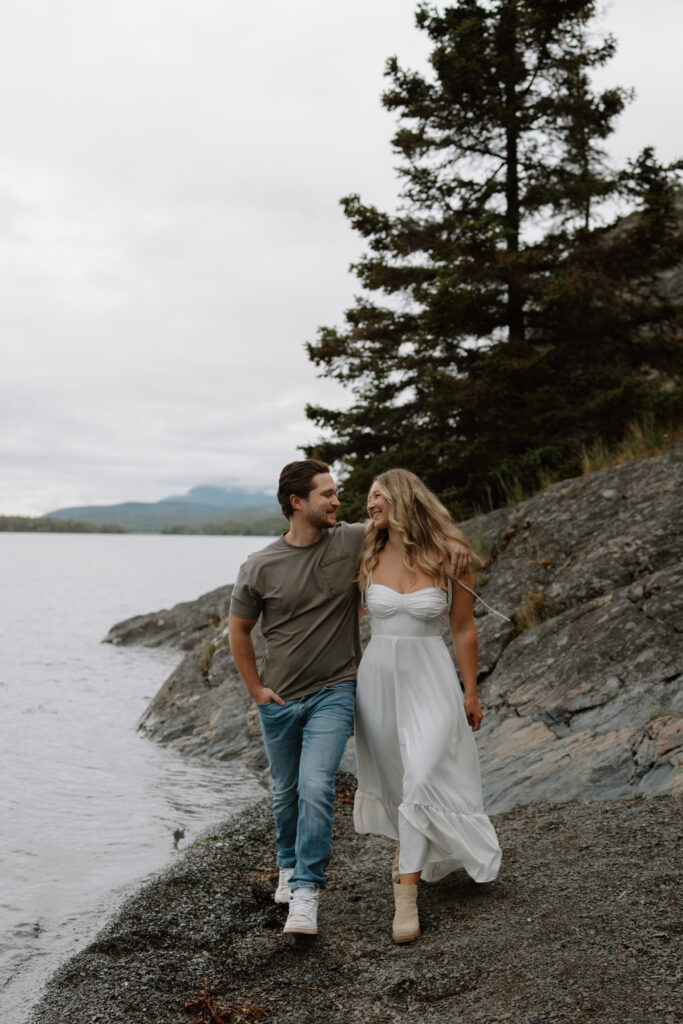 Engaged couple walking along the shoreline of Kenai Lake in Cooper Landing Alaska with arms wrapped around each other's waist