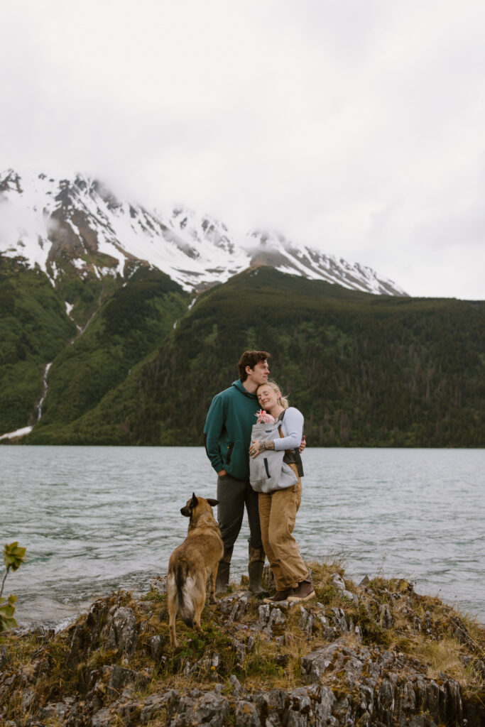 Family standing on the rocky edge of Kenai Lake Alaska, mom wearing baby in a chest carrier with dad and dog by their side, mountains behind