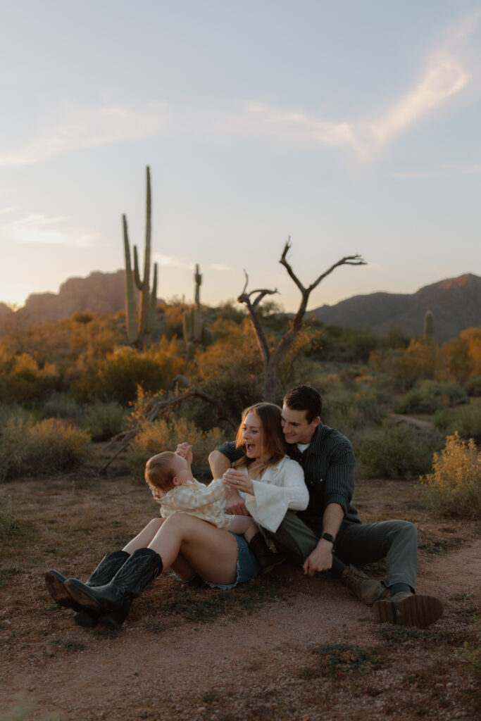 Family sits on the desert ground. Daughter is on moms holding each other's hands. They are smiling and it is sunset.