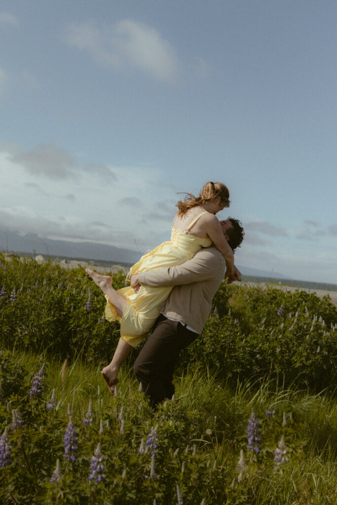 Couple in Homer Alaska, boyfriend picking up girlfriend as she wraps her arms around his neck and pops one foot into the air surrounded by grass and wild lupine flowers