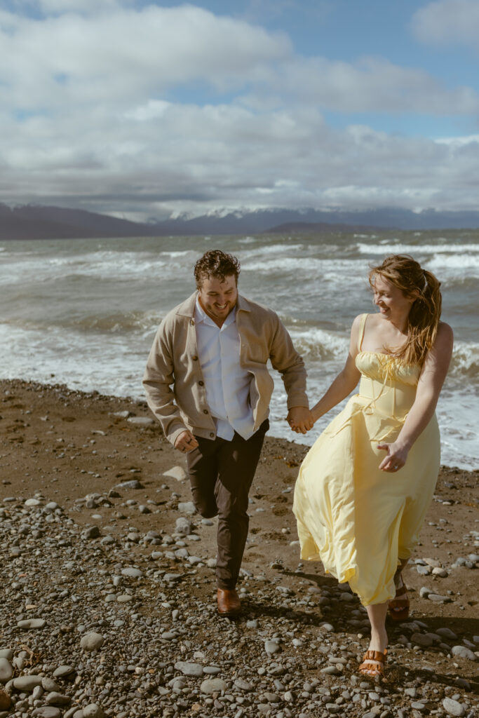 Couple running up the beach hand in hand in Homer Alaska smiling as waves come in behind them