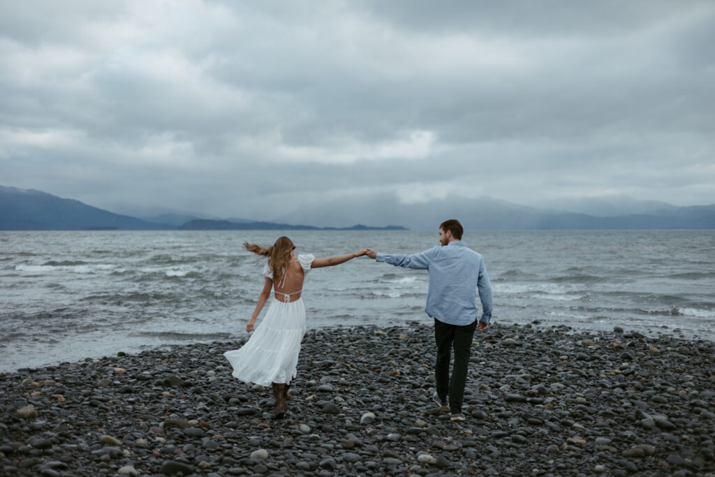Couple walks towards ocean on an overcast day as they hold hands in Homer Alaska