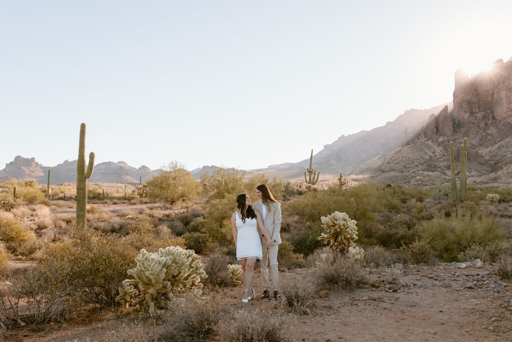Engaged couple at Lost Dutchman State Park, Arizona. Man stands behind woman as she looks over her shoulder at him, with the sun rising over the Superstition Mountains in the background during golden hour engagement session.
