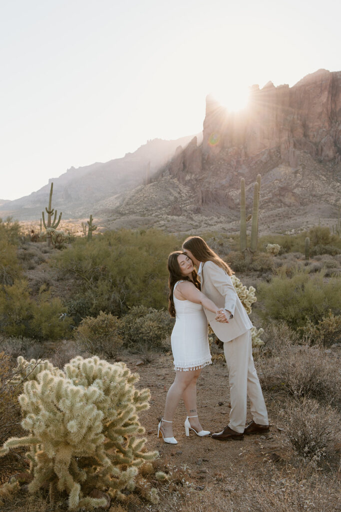 Engaged couple at Lost Dutchman State Park, Arizona. Couple face each other and have hands interlaced as man kisses face of woman, with the sun rising over the Superstition Mountains in the background during golden hour engagement session.