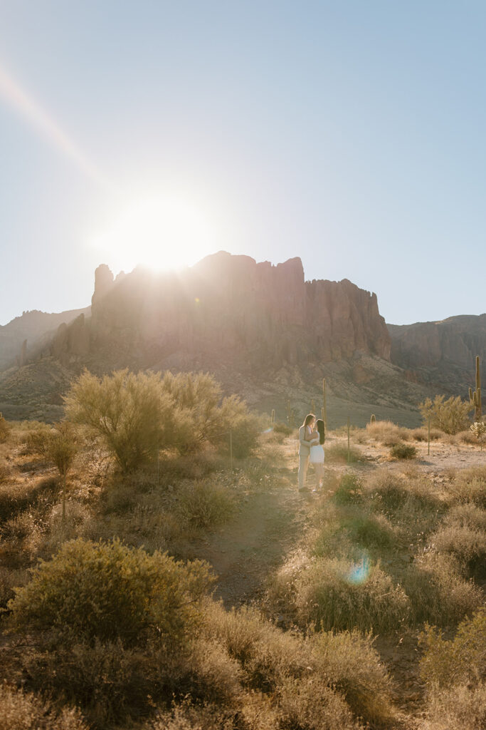 Engaged couple at Lost Dutchman State Park, Arizona. Couple faces towards each other, with the sun rising over the Superstition Mountains in the background during golden hour engagement session.