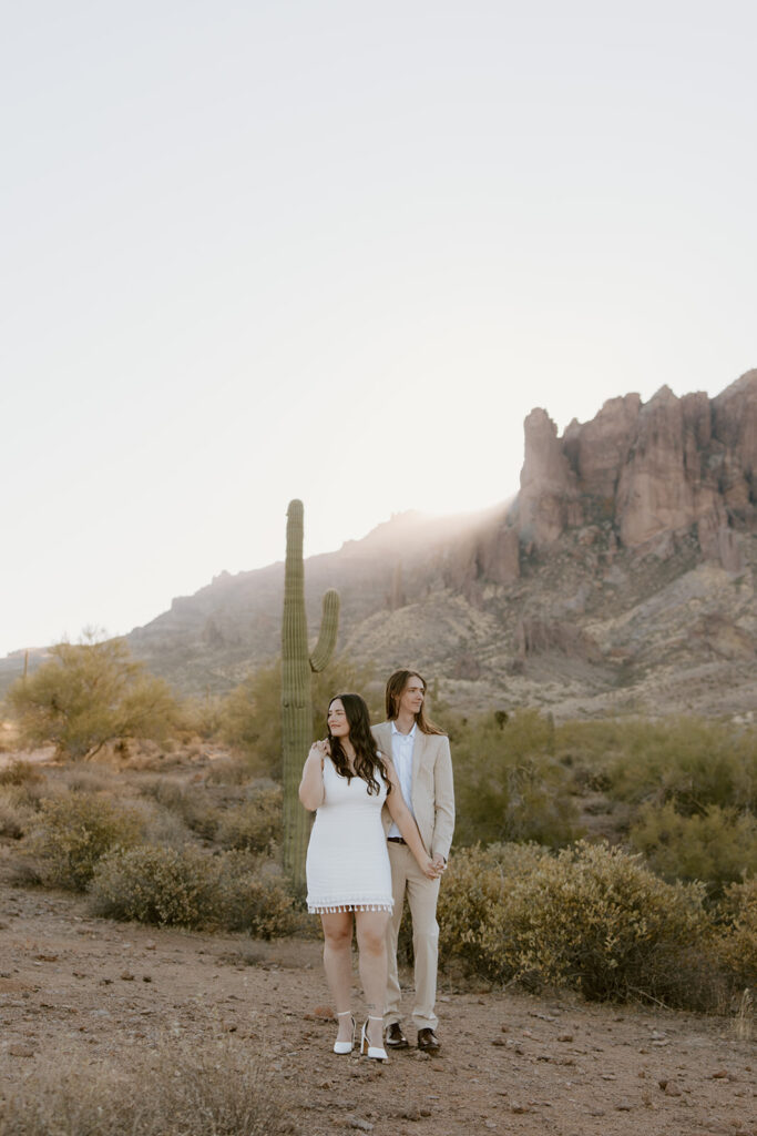 Engaged couple at Lost Dutchman State Park, Arizona. Man stands behind woman as they look opposite directions, with the sun rising over the Superstition Mountains in the background during golden hour engagement session.