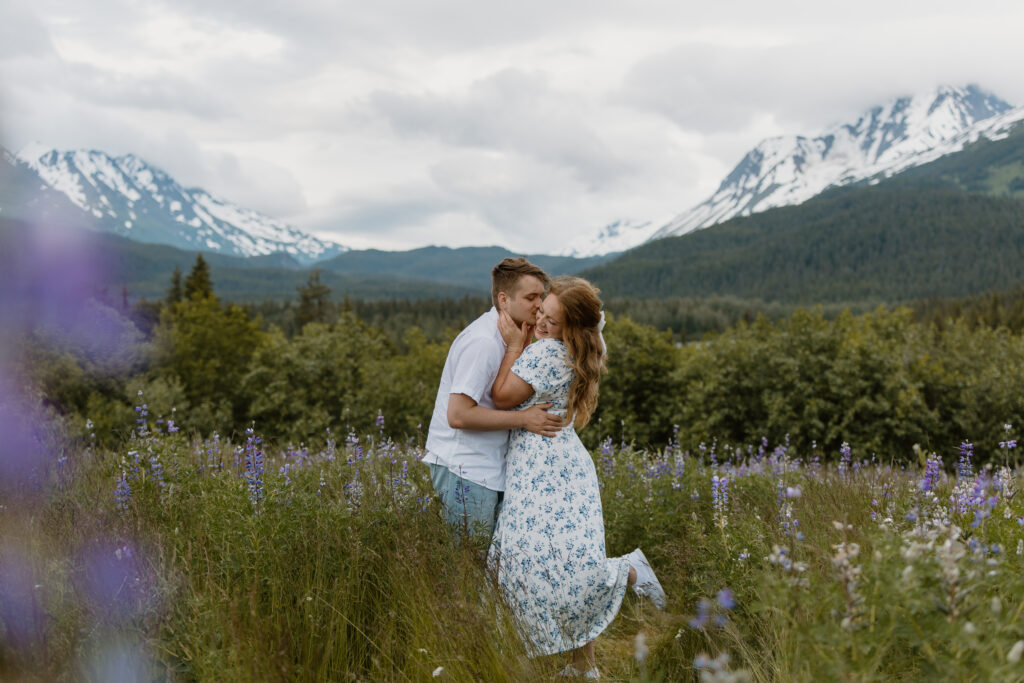 A couple embraces as the boyfriend kisses his girlfriend's cheek. They are standing in a field of lupine with mountains behind them.